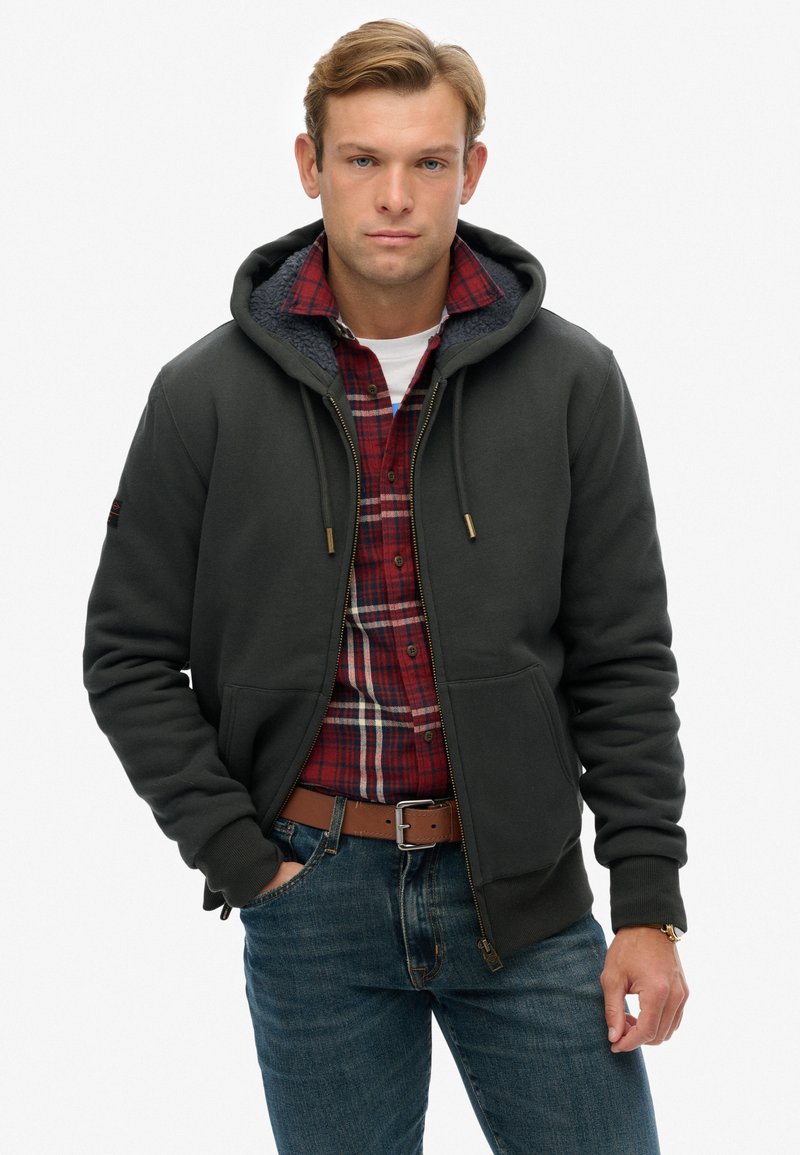 Sudadera con capucha en gris oscuro, con cremallera frontal y bolsillos tipo canguro, llevada sobre una camisa de cuadros roja, combinada con jeans azules.