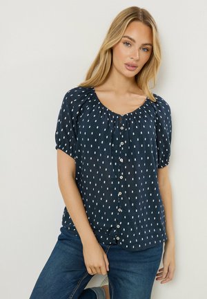 Blonde vrouw in een marineblauw blouse met korte mouwen en witte stippen, gecombineerd met een blauwe spijkerbroek, staand tegen een witte muur.
