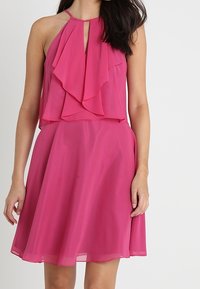 Fuchsia halterneck-klänning med lager av volanger på bodice, nyckelhål-detalj och en utsvängd kjol, tillverkad av lätt och mjukt tyg.