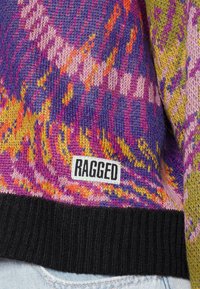 Wielokolorowy sweter z dzianiny z abstrakcyjnymi wzorami w odcieniach fioletu, żółci, pomarańczu oraz czarnym ściągaczem, oznaczony etykietą "RAGGED", noszony na jasnoniebieskich dżinsach.