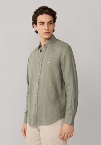 Homme portant une chemise à manches longues vert clair boutonnée avec col et un pantalon beige, regardant sur le côté sur un fond uni.