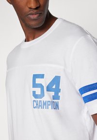 Tricou alb cu mânecă scurtă, prezentând numerele albastre imprimate "54" și "CHAMPION" cu un design texturat și accente albastre în dungi pe mâneci.