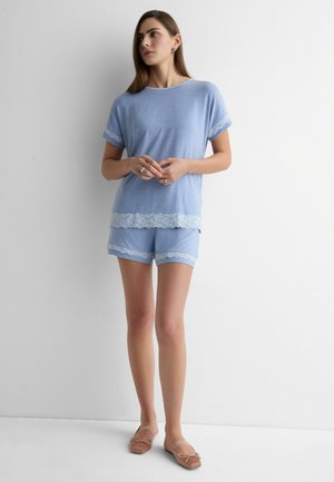 Femme debout contre un mur, portant un haut à manches courtes bleu clair et un short assorti avec une bordure en dentelle blanche, ainsi que des ballerines marron.