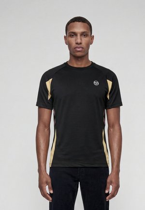 Homme portant une chemise noire à manches courtes avec des panneaux latéraux beiges et un petit logo blanc sur la poitrine, debout devant un fond uni.