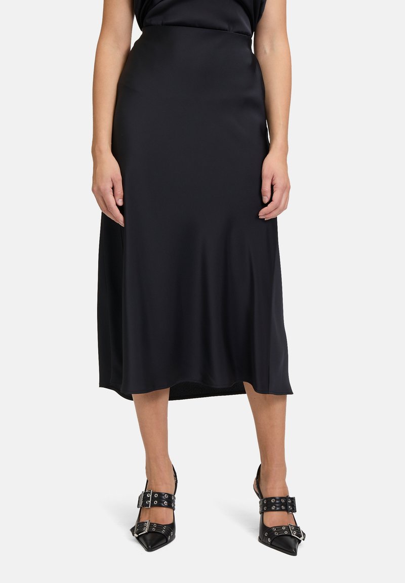 Schwarzer Satin-Midirock mit glatter Textur, hoher Taille und leicht ausgestelltem Saum. Kombiniert mit schwarzen spitzen Schuhen.