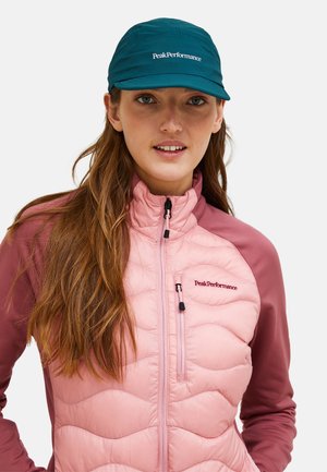 Woman wearing teal PeakPerformance cap and pink zip-up quilted jacket with maroon sleeves, facing forward with neutral expression.