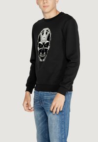 Sudadera negra con un diseño gráfico de calavera que presenta ojos verdes, cuello redondo y puños de canalé. Combinada con jeans de color azul claro.