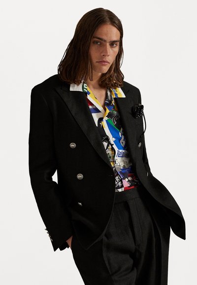 Blazer negro de doble botonadura con botones plateados sobre una camisa de estampado colorido. Presenta un pañuelo de bolsillo y pantalones ajustados.