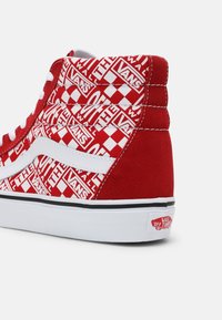 Baskets montantes Vans rouges et blanches avec motif damier et logos "Vans Off The Wall" sur des panneaux en tissu et semelle en caoutchouc blanche.