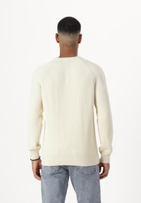 Rippensweater in Creme mit rundem Halsausschnitt und langen Ärmeln, der einen lockeren Schnitt und eine strukturierte Oberfläche aufweist. Kombiniert mit hellblauer Denim.