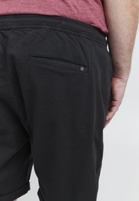 Gros plan sur une personne portant un short noir avec une poche arrière et un petit logo rond, associé à un t-shirt bordeaux.