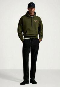 Hoodie vert olive avec une poche avant et un logo, associé à un pantalon noir fuselé et des baskets noires. Une casquette de baseball sombre complète la tenue.