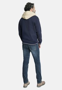 Sudadera marina con capucha beige, puños y dobladillo acanalados, combinada con unos vaqueros de mezclilla azules y zapatos marrón claro. Persona vista desde atrás.