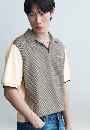 Νέος άνδρας που φοράει μπλούζα πόλο με ασπρόμαυρο καρό και κρεμ μανίκια, καθώς και μπλε τζιν, κοιτάζει προς τα αριστερά με το ένα χέρι στην τσέπη.