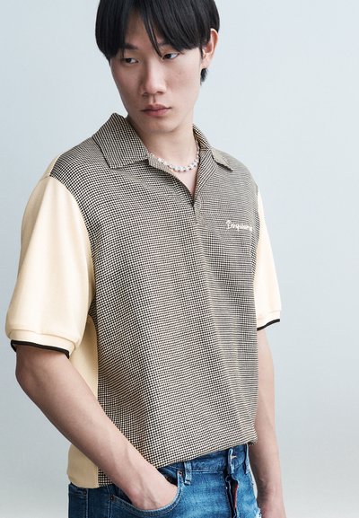 Jeune homme portant un polo à carreaux noir et blanc avec des manches couleur crème et un jean bleu, regardant vers sa gauche avec une main dans la poche.