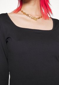 Person med klart röd hår som bär en svart topp med fyrkantig halsringning och ett kraftigt guldhalsband med en lås.