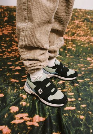 Person wearing beige corduroy pants and black and gray sneakers with velcro straps standing on floral patterned ground.