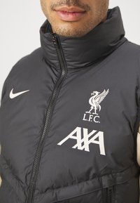 Gros plan d'une personne portant une veste noire zippée avec le logo de Liverpool FC, le sponsor AXA et le swoosh Nike à l'avant.