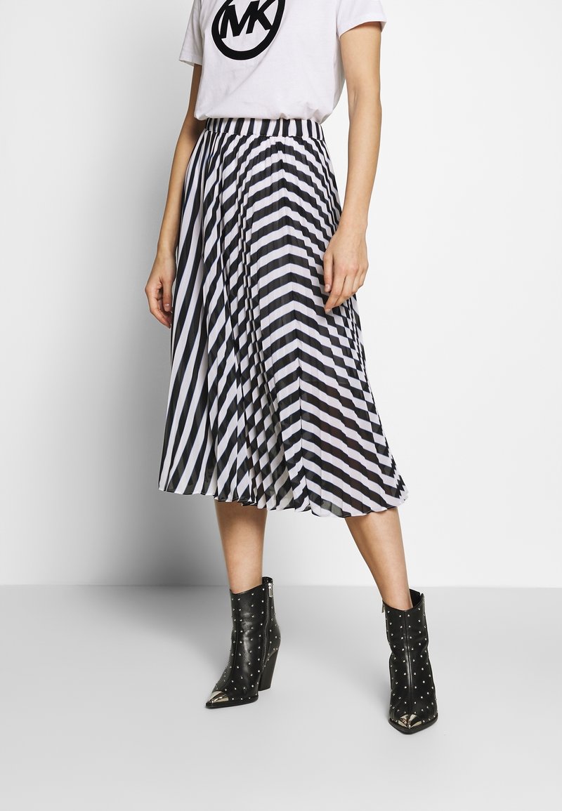 Kvinna iklädd en svartvit diagonalt randig plisserad midikjol, vit t-shirt och svarta spikade ankelboots med spetsiga metalltå.