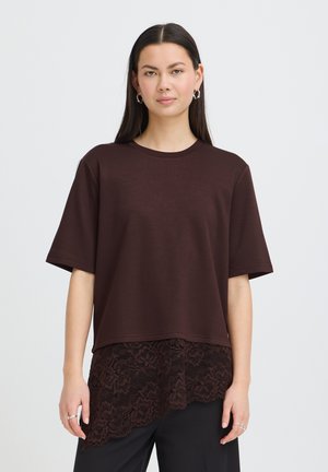 Femme aux longs cheveux foncés porte un haut marron ample à manches courtes avec un ourlet asymétrique en dentelle, un pantalon noir, des boucles d'oreilles créoles argentées et des bagues.