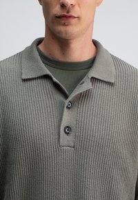 Lichtgrijze geribbelde polo met een kraag, voorzien van drie donkere knopen, gedragen over een donkergroene ronde hals t-shirt.