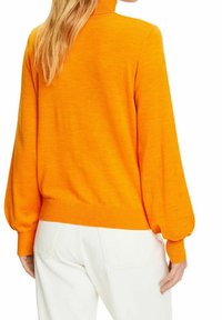 Pull en tricot orange vif avec un col montant, des manches longues bouffantes et une taille cintrée, associé à un pantalon blanc, présentant une texture lisse.