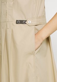 Beigenvärinen mekko, jossa rento istuvuus. Siinä on musta kangasvyö, jossa on hopeanväriset silmukat ja nappi, sekä näkyvä sivutasku.