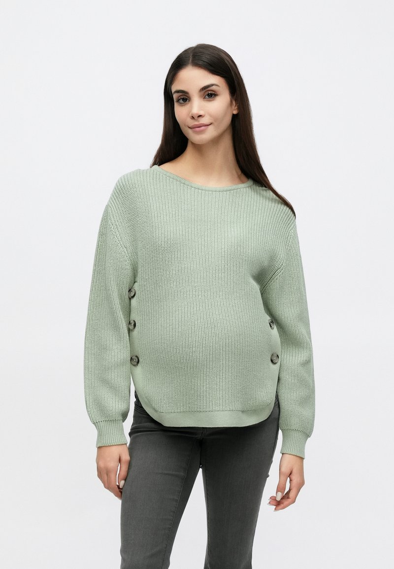 Pull tricot vert clair avec une texture côtelée, un col large et des boutons décoratifs sur le côté. Associé à un pantalon gris.