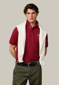 Červená polo tričko s dvoma gombíkmi a vyšitým logom, doplnené olivovozelenými nohavicami a bielym sveterom prehodeným cez plecia.