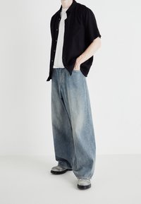 Camisa negra de manga corta con botones, camiseta blanca debajo, combinada con pantalones vaqueros de pierna ancha en denim azul claro y calzado gris. Fondo simple.