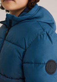 Mėlyna pamušta striukė su gobtuvu, su juoda užtrauktuku ir apvaliu juodu logotipo ženklu ant rankovės. Sklandi tekstūra, siuvinėjimo dizainas.