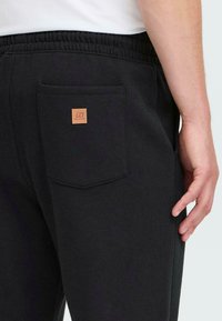 Nahaufnahme einer Person in schwarzen Jogginghosen mit elastischem Bund und einer Gesäßtasche, die mit einem kleinen braunen Markenpatch versehen ist.