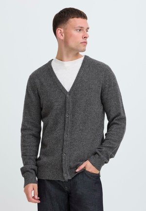 Grå strikket cardigan med v-hals, frontknapper og ribbede mansjetter. Brukt over en hvit skjorte, vist mot en lys bakgrunn.