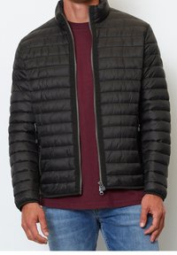 Mand iført en sort quiltet jakke over en maroon skjorte og blå jeans, med jakken delvist åbnet.