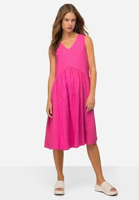 Abito di lino rosa con scollo a V, senza maniche e un silhouette a strati e fluida. Abbinato a scarpe slip-on di colore chiaro.