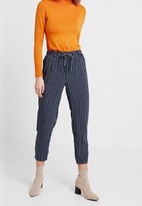 Granatowe spodnie w paski z elastycznym paskiem, dopasowany krój i zwężane nogawki, zestawione z dopasowanym pomarańczowym golfem i beżowymi botkami za kostkę.