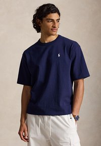 Homme aux cheveux bouclés et foncés portant une chemise bleu marine à manches courtes avec un logo et un pantalon blanc à cordon, regardant sur le côté.