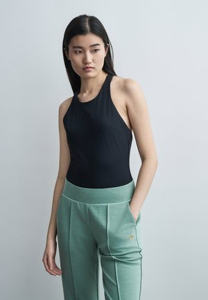 Ung kvinde iført en sort ærmeløs top og mintgrønne bukser med hænderne i lommerne, kiggende til siden mod en ensartet baggrund.