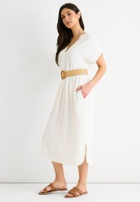 Abito midi bianco con maniche corte, dotato di scollatura a V, una cintura intrecciata beige in vita e tasche laterali. Sandali piatti marroni.