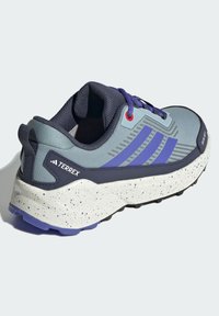 Vīriešu sporta apavi ar pelēku un jūras zila augšdaļu, trīs violetām svītrām, sarkaniem acs caurumiem un plankumainu baltu zoli, kas nodrošina saķeri.