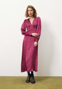 Robe longue en satin prune avec un décolleté en V, un bouton à l'avant, des manches longues bouffantes et une silhouette cintrée. Associée à des sandales à talons noires.