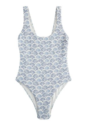 Femeie purtând un costum de baie albastru și alb cu model de scoici marine, stând pe o plajă cu nisip lângă stânci și un prosop roșu în dungi.