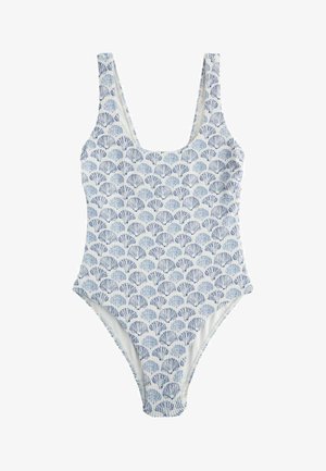 Femme portant un maillot de bain bleu et blanc à motif de coquillages, debout sur une plage de sable près de rochers et une serviette rouge à rayures.