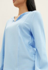 Lys blå bluse med vertikale hvite striper, avslappet passform, V-hals og lange ermer. Stoffet ser jevnt og noe teksturert ut.