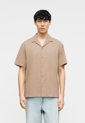 Mężczyzna ubrany w beżową koszulę na krótki rękaw z zapinanym kołnierzem typu camp oraz jasnoniebieskie dżinsy, stojący na jednolitym białym tle.