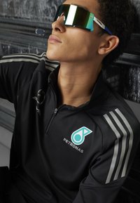 Sports solbriller med grønfarvede spejlede linser, hvide og blå detaljer. Modellen har en sort zip-up jakke på med sølvstriber og logo.