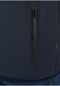 Gros plan d'une poche de veste bleu marine avec une fermeture éclair noire verticale et un cordon noir et blanc, portée sur un jean bleu.