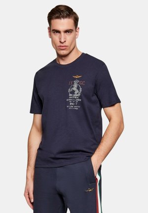 Uomo che indossa una maglietta blu navy con stemma e testo, abbinata a pantaloni blu navy con strisce rosse, bianche e verdi sul lato.