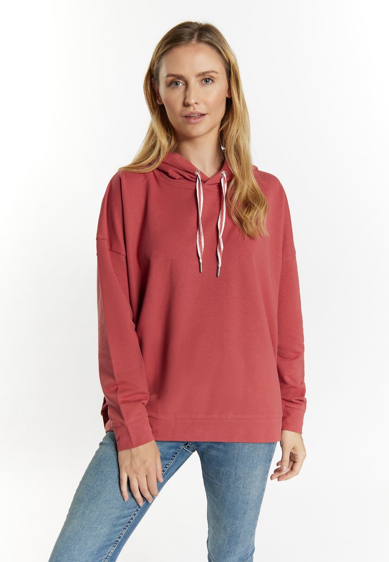 Felpa rossa con una texture morbida, chiusura con coulisse e aperture laterali. Indossata con jeans blu, caratterizzati da un fit rilassato e oversize.