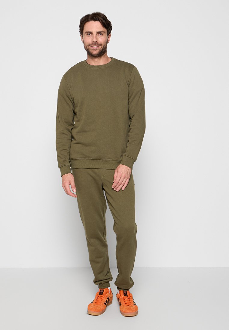 Sweatshirt vert olive et sweat-pants assortis en tissu doux et texturé. Portés avec des baskets orange vif, se tenant devant un fond uni.